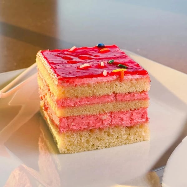 Layered vanilla cake with pink frosting and red jelly topping with colorful sprinkles on a white plate.