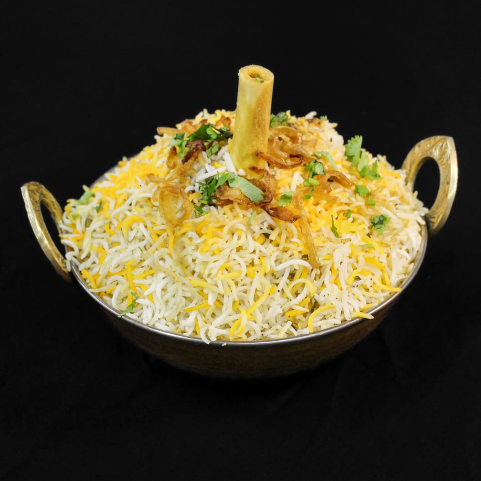 Little Spice Indian Cuisine's lamb shank biryani, garnished with fried onions and cilantro in a brass bowl.