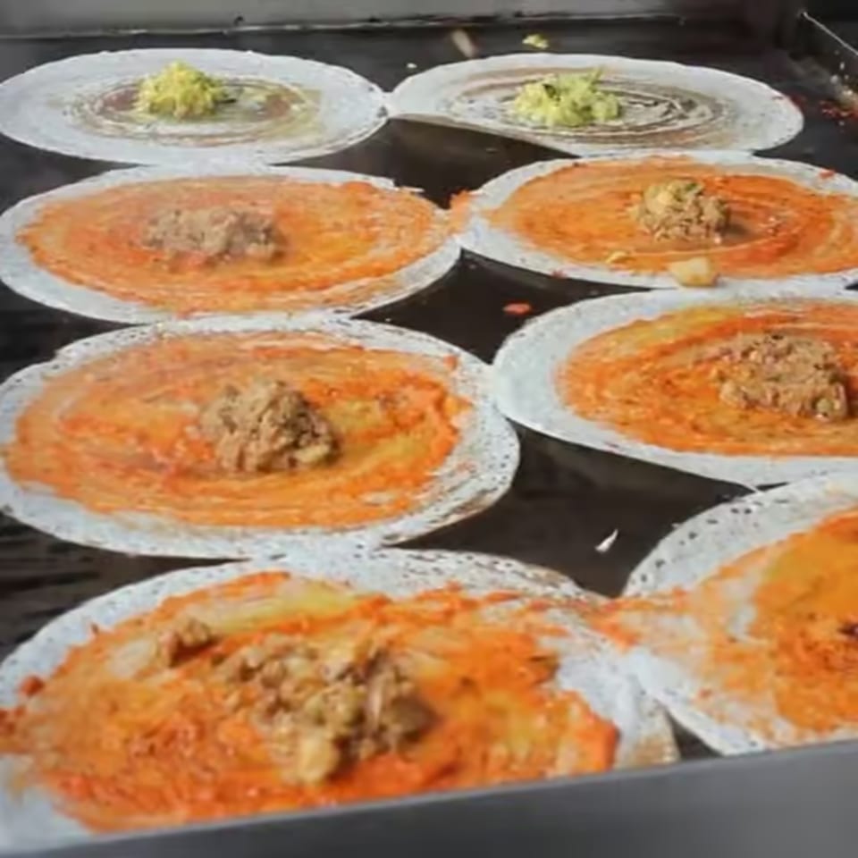 Multiple dosas cooking on a griddle, some with green filling, others with orange sauce and brown filling.