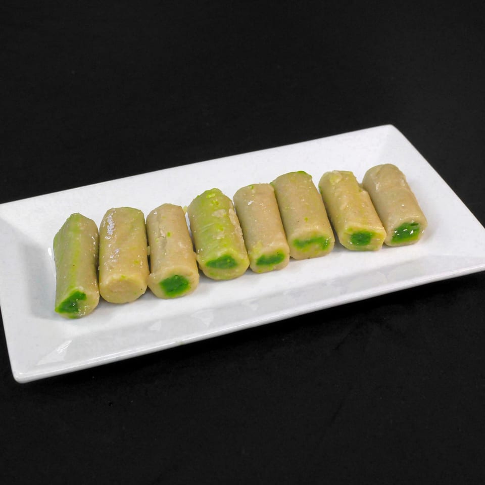 Eight Indian sweets, light brown with green centers, arranged on a white rectangular plate.
