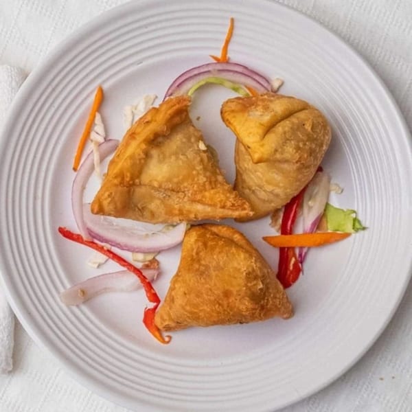 Three golden-brown samosas on a white plate, garnished with red onion, carrots, and red chili.