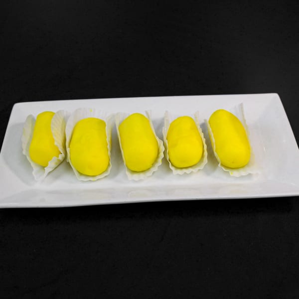 Five bright yellow Indian sweets, possibly mango barfi, in white paper cups on a white rectangular plate.