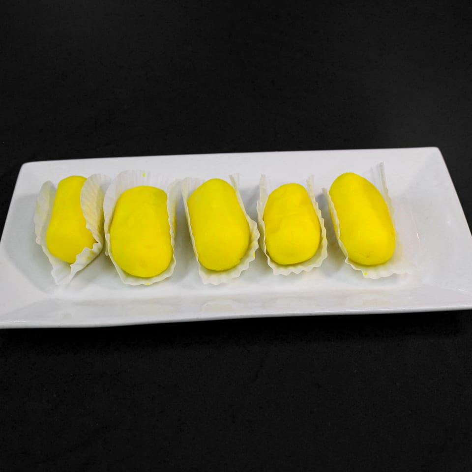 Five bright yellow Indian sweets, possibly mango barfi, in white paper cups on a white rectangular plate.