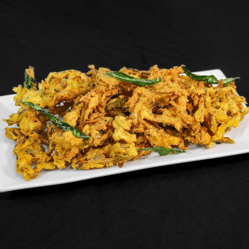 A white rectangular plate filled with golden-brown, crispy onion bhaji, garnished with fresh green curry leaves.