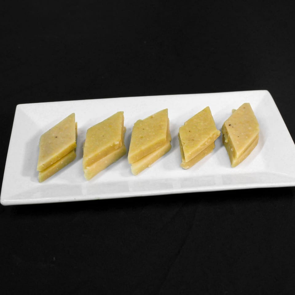 Five diamond-shaped pieces of light yellow Indian barfi dessert on a white rectangular plate.