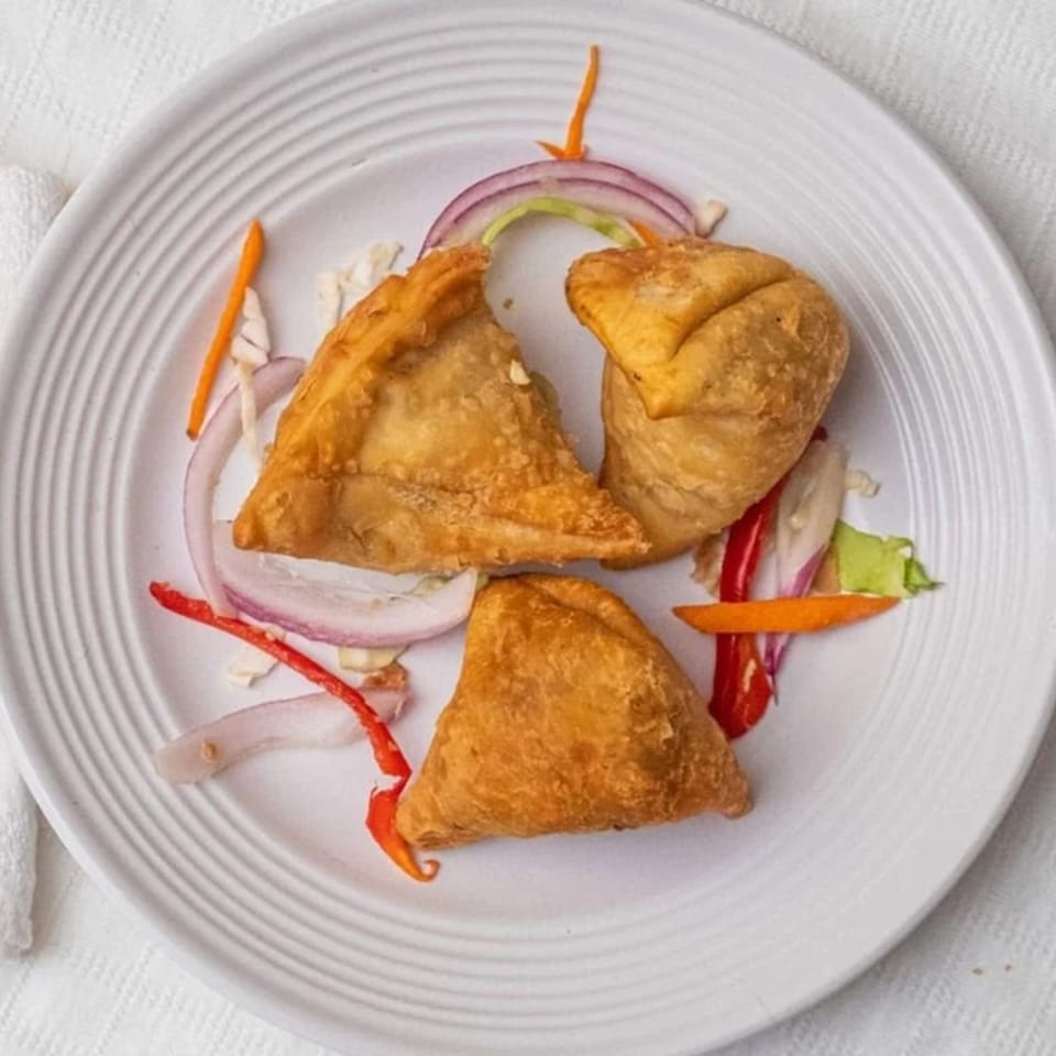Three golden-brown samosas on a white plate, garnished with sliced red onions, carrots, and peppers.